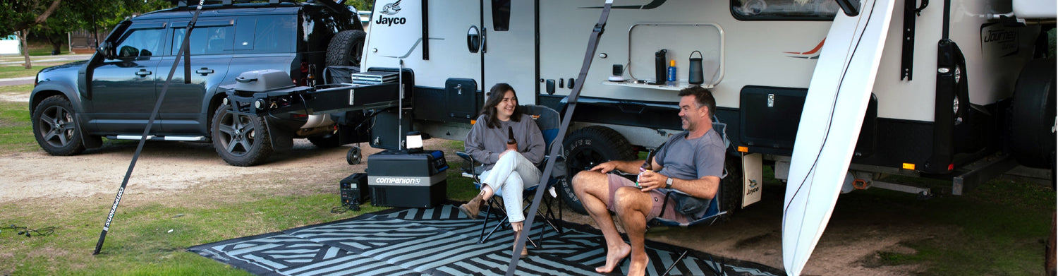 A couple sitting outside a camper, smiling and relaxing with drinks, near a parked SUV and bike.